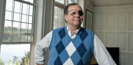 Portrait of Peter L. in his home, wearing a sweater vest, and a black eye patch under his glasses