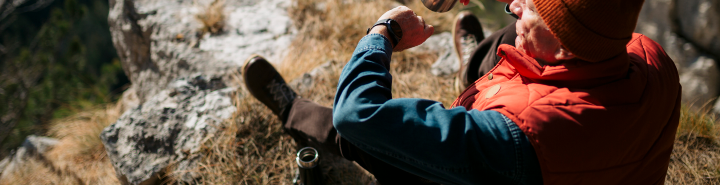 Man sitting on a mountainside looking at his watch holding an empty cup